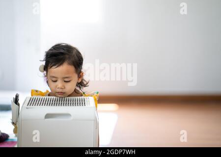 Photo d'une petite fille asiatique debout près de l'air conditionné portable et curieusement la regardant Banque D'Images