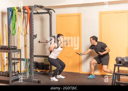 Sportswoman faisant des squats avec bar avec entraîneur dans la salle de gym Banque D'Images