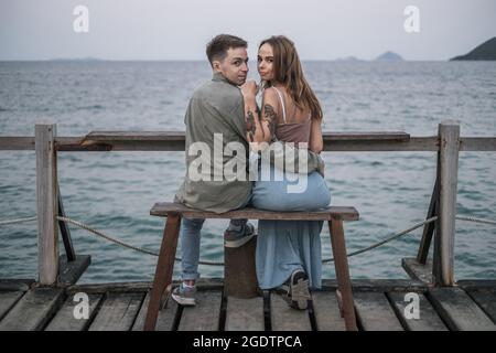 Beau jeune couple amoureux assis sur le banc au bord de la mer, en cuddling et regardant l'appareil photo avec l'espace de copie. Photo de haute qualité Banque D'Images