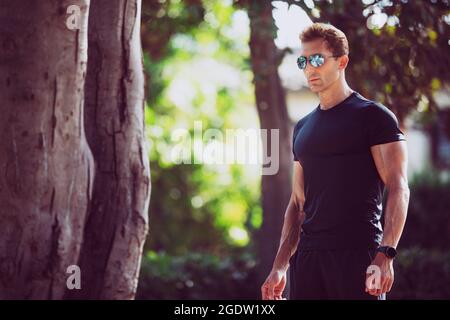 Homme sportif avec lunettes de soleil dans un parc extérieur. Il porte un maillot de sport, un look sérieux. Banque D'Images