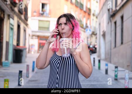 Jeune femme caucasienne avec des cheveux roses frognant tout en parlant sur le smartphone et de l'eau potable dans la rue. Banque D'Images