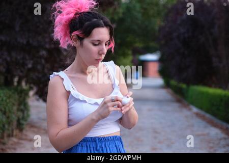 Jeune femme caucasienne aux cheveux roses mettant des oreillettes sans fil dans un parc. Banque D'Images