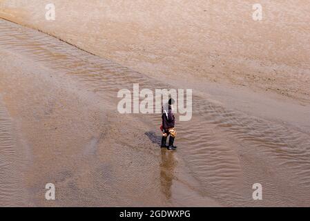 Femme en chapeau et bien-être regardant vers le bas en marchant sur une plage de sable à travers un ruisseau d'eau Banque D'Images