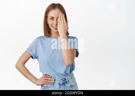 Belle femme blonde couvre la moitié du visage, regardant avec un oeil et souriant confiant, avant et après effet, debout dans le bleu t-shirt contre blanc Banque D'Images