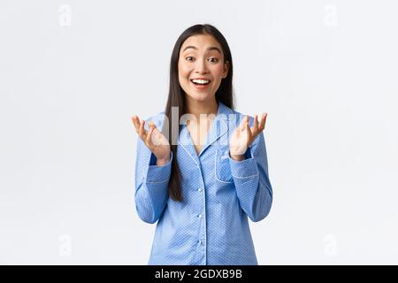 Bonne fille asiatique en pyjama bleu levant les mains et applaudir dans le plaisir, souriant émerveillé, écoutant de merveilleuses nouvelles, louant le bon travail Banque D'Images