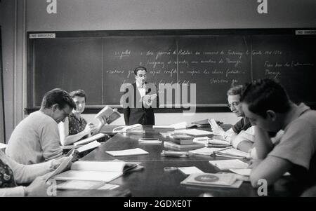 1950s, historique, professeur d'université adulte, habillé avec intelligence d'une veste et d'un noeud papillon, debout devant un tableau noir, enseignant la littérature française à un groupe de étudiants assis autour d'une table avec leurs notes et livres, des Fables, Iowa, États-Unis. Banque D'Images