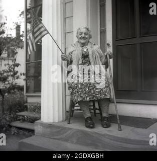 Années 1950, historique, une fière américaine... une dame âgée assise sur une chaise en bois sur un pas en pierre devant sa porte d'entrée, avec une baguette dans une main et dans l'autre, le drapeau américain, États-Unis. Avec ses 50 étoiles et ses 13 bandes, le drapeau est le symbole puissant de la nation américaine, Banque D'Images