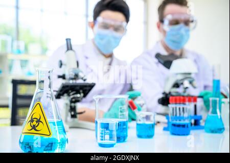 Équipement de laboratoire avec liquide bleu à l'intérieur du support sur la table avec couple scientifique mâle portant une combinaison de protection travaillant avec un microscope pour la recherche à l Banque D'Images