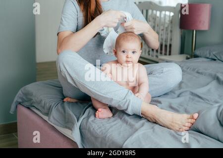 Une jeune mère est assise sur le lit et habille son bébé Banque D'Images