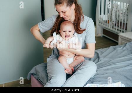 Une jeune mère habille son bébé gai assis sur ses genoux et montrant la langue. Banque D'Images