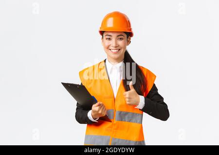 Satisfaite femme asiatique ingénieur de construction satisfait des résultats de l'inspection, portant des vêtements de sécurité et un casque dans la zone de construction, écrit Banque D'Images