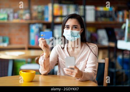 Bonne femme latine portant un masque, se reposant dans un café et faisant le paiement par carte de crédit et smartphone Banque D'Images