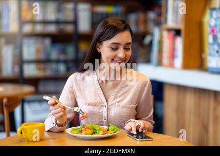 Bonne jeune femme arabe envoyant un SMS sur un smartphone tout en mangeant de la salade dans un café, en utilisant une application pour envoyer un sms Banque D'Images