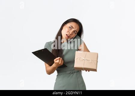 Occupé confus femme asiatique répondant à un appel téléphonique, parlant avec le client perplexe, tenant la boîte de livraison, préparer le paquet pour l'expédition, l'enregistrement avec Banque D'Images