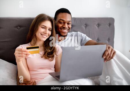 Couple interracial gai avec carte de crédit et achat d'ordinateur portable en ligne ensemble, faisant le paiement sans contact sur le lit Banque D'Images
