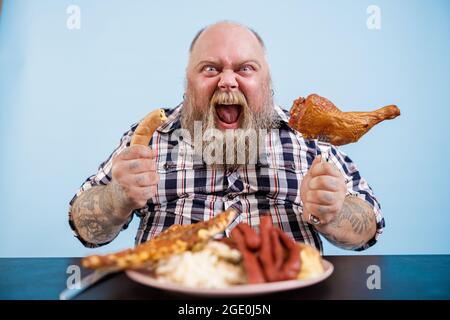 L'homme obèse émotif tient la saucisse et la jambe de poulet fumé assis à la table Banque D'Images