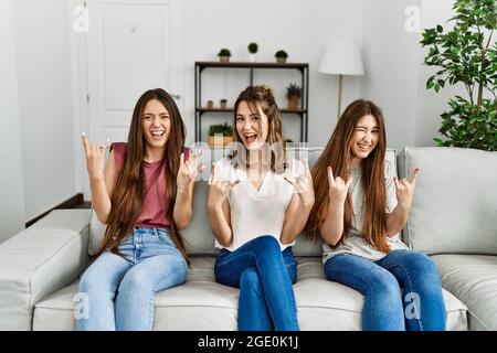 Groupe de trois filles hispaniques assis sur le canapé à la maison criant avec l'expression folle faisant le symbole de rock avec les mains vers le haut. Star de la musique. Concept lourd. Banque D'Images