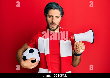 Jeune homme hispanique football hooligan tenant le ballon en utilisant le mégaphone dans le visage de choc, regardant sceptique et sarcastique, surpris par la bouche ouverte Banque D'Images