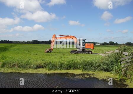 Pelle hydraulique rouge orangée néerlandaise dans un paysage de prairie avec des arbres, des fossés et des dunes au loin. Été, août, pays-Bas Banque D'Images