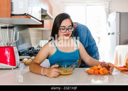 Jeune fille latine avec le syndrome de Down manger des fruits dans la cuisine, dans le concept de handicap en Amérique latine Banque D'Images