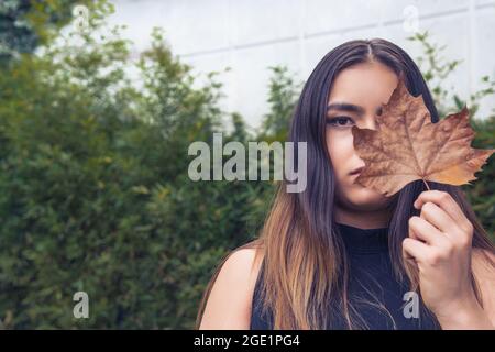 Portrait horizontal d'une adolescente Latina à poil droit couvrant un œil avec une feuille d'érable Banque D'Images