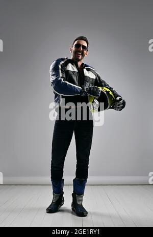 Heureux non rasé homme adulte motocross motard coureur en moto cuir équipement bottes, veste et gants marche avec son casque Banque D'Images