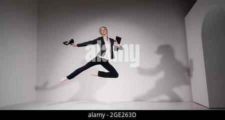 Une ballerine pieds nus gracieuse dans un costume d'affaires sautant avec des chaussures dans ses mains sur un fond blanc. Banque D'Images