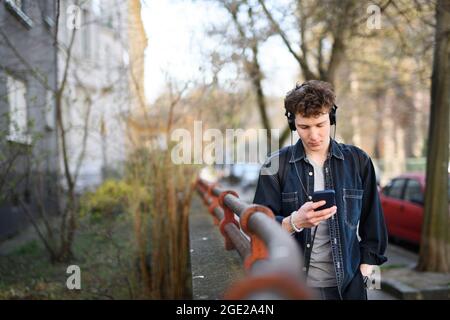 Portrait d'un jeune homme en déplacement avec un casque qui marche à l'extérieur en ville, à l'aide d'un smartphone. Banque D'Images