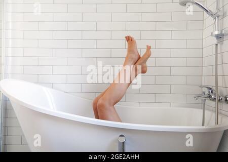 Femme pieds nus inreconnaissables dans la baignoire et se levant les jambes contre un mur de carrelage blanc pendant les soins de la peau dans la salle de bains à la maison Banque D'Images