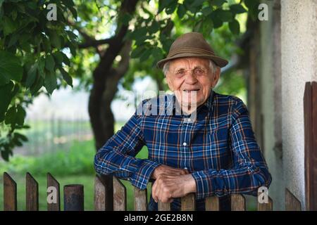 Portrait d'un homme âgé debout à l'extérieur dans le jardin, penché sur une clôture en bois et regardant l'appareil photo. Banque D'Images