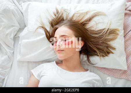 La jeune femme est endormie avec les yeux fermés applique des patches au visage après le sommeil. Soins de la peau matinaux taches oculaires d'hydrogel au lit Banque D'Images