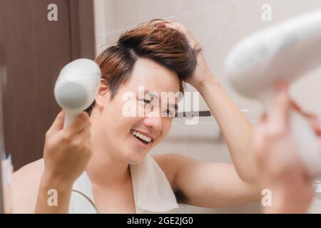 Un jeune homme tendance séchant ses cheveux avec un sèche-cheveux devant le miroir de salle de bains Banque D'Images