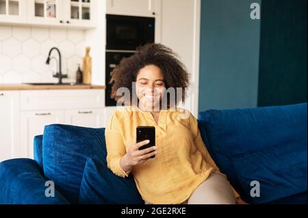 Joyeuse femme afro-américaine utilisant un smartphone assis sur le canapé dans un salon confortable et élégant, bavardant et envoyant des messages en ligne, passant des loisirs sur les réseaux sociaux, partageant des informations avec des amis Banque D'Images