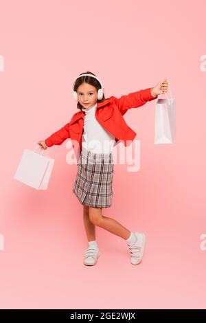 fille en blouson orange, jupe à carreaux et casque posant avec des sacs d'achats sur rose Banque D'Images