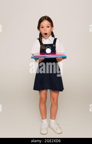 fille stupéfait dans l'uniforme de l'école tenant des cahiers et petit réveil sur gris Banque D'Images