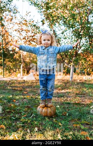 Petite fille se tient sur la grande citrouille orange avec ses bras écartés jouer dehors avec la récolte d'automne. Jour de Thanksgiving Banque D'Images