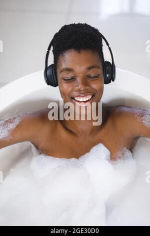 Femme afro-américaine souriante se détendant dans le bain avec les yeux fermés en écoutant de la musique sur les écouteurs Banque D'Images