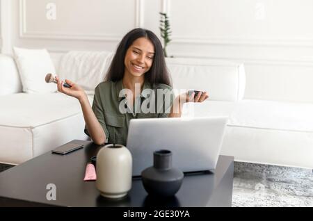 Femme indienne souriante appliquant de la poudre avec le pinceau et regardant des didacticiels de maquillage sur ordinateur portable, fille orientale souriante apprenant à faire le maquillage par elle-même avec un tuteur en ligne. Concept de passe-temps Banque D'Images