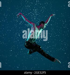 Un jeune danseur de ballet masculin en mouvement, action sous la pluie sur fond bleu à la lumière du néon. Banque D'Images