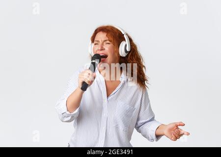 Emotions, style de vie et loisirs. Insouciante passionnée redhead femme d'âge moyen appréciant chanter émotionnellement karaoké, tenant le microphone et Banque D'Images