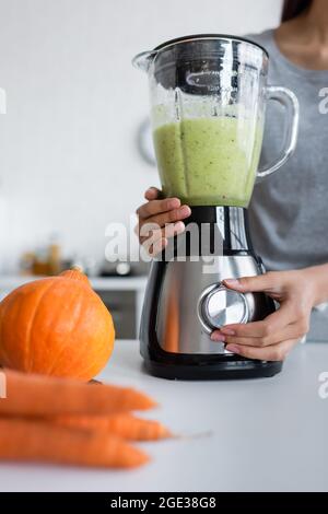 vue rognée d'une femme préparant un smoothie végétal près de la citrouille et des carottes floues Banque D'Images