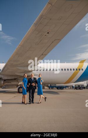 Vue en longueur d'un pilote debout avec deux hôtesses en uniforme bleu clair devant un avion par temps ensoleillé Banque D'Images