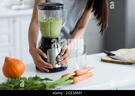 vue partielle de la femme préparant du smoothie frais près de la citrouille et des carottes sur la table de cuisine Banque D'Images