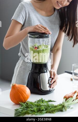 vue partielle d'une femme asiatique souriante préparant un smoothie près de la citrouille crue et des carottes Banque D'Images