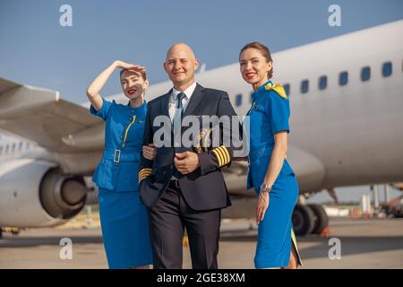 Heureux pilote et deux hôtesses attrayantes debout ensemble devant un avion et souriant après l'atterrissage Banque D'Images