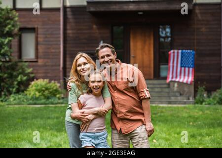 Une famille souriante se hante près d'un drapeau américain flou sur la maison à l'extérieur Banque D'Images