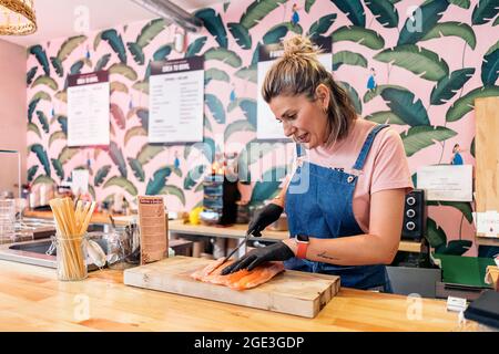 Femme employée de restaurant portant un tablier et des gants en latex coupant du saumon pour un poke Bowl. Banque D'Images