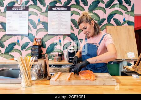 Joli employé de restaurant portant un tablier et des gants en latex coupant du saumon pour un poke Bowl. Banque D'Images