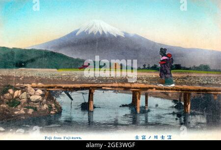 Ancienne carte postale japonaise de couleur main d'une femme de kimono traversant une petite passerelle en bois avec le Mont Fuji en arrière-plan. Vers 1910 Banque D'Images