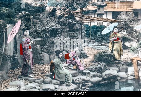 Vieille carte postale japonaise colorée à la main vers 1900, de quatre femmes Geisha sur une rive avec l'une d'entre elles pêchant et deux autres tenant des parasols. Banque D'Images